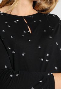 Zwarte lange mouwen top met een keyhole-halslijn, uitgerust met een borduursel van witte sterrenpatronen. Zacht materiaal en een relaxte pasvorm.