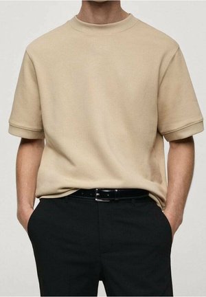 Beige kurzärmliges Sweatshirt mit geripptem Rundhalsausschnitt, umgeschlagenen Bündchen und lockerem Schnitt, kombiniert mit einer schwarzen Hose und einem schwarzen Gürtel.