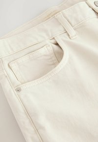 Beige denim jeans med en frontlomme med syningsdetaljer, metalnitteaccent og subtil stribetekstur.