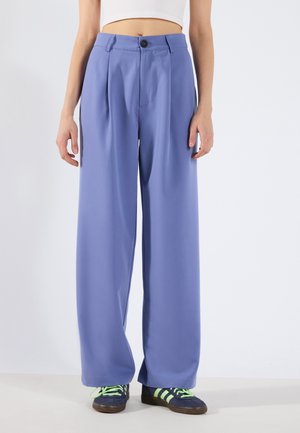 Femme portant un pantalon ample taille haute lavande, un top court blanc et des baskets bleu marine avec des rayures et lacets vert citron, debout sur un sol blanc.
