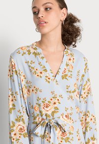 Vestido azul claro com estampa floral em pêssego e verde. Apresenta um decote em V, mangaslongas e uma cintura atada para um estilo ajustado.