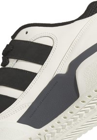 Scarpa sportiva bianca con accenti neri, caratterizzata da tre strisce nere, sovrapposizioni testurizzate e una suola in gomma con grip a motivi.