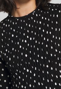 Czarna bluzka z teksturowanej tkaniny w białe kropki, z pomarszczoną powierzchnią i okrągłym dekoltem. Zbliżenie na detale.
