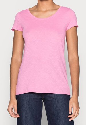 Kobieta ubrana w jednokolorowy różowy T-shirt z krótkim rękawem i ciemnoniebieskie dżinsy, stojąca z rozluźnionymi rękami wzdłuż boków na jednolitym tle.