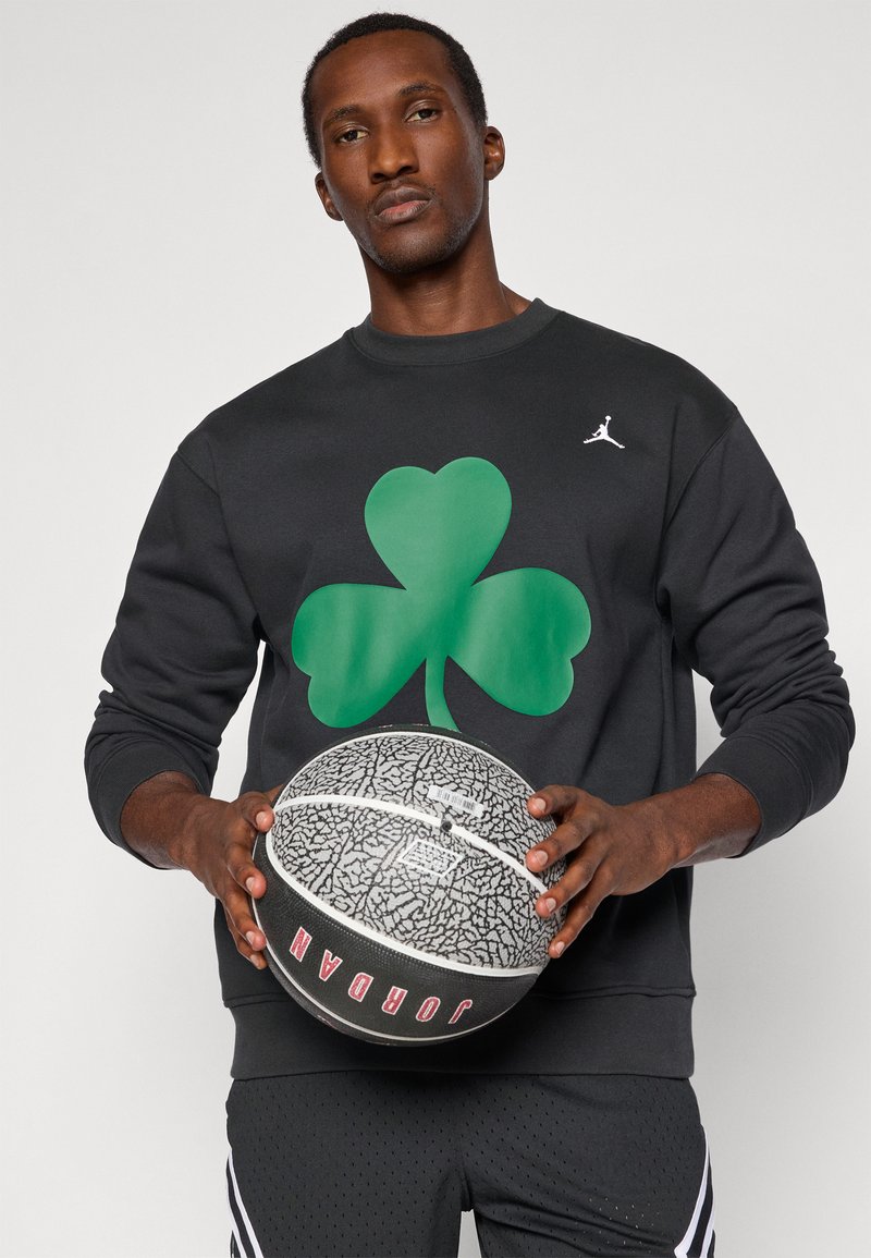 Man holding a black and gray basketball, wearing a black sweatshirt with a large green shamrock and a small white Jumpman logo.
