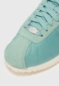 Gros plan sur la pointe d'une basket bleu sarcelle avec tissu en daim et nylon, lacets texturés et petite étiquette argentée avec le logo Nike sur les lacets.