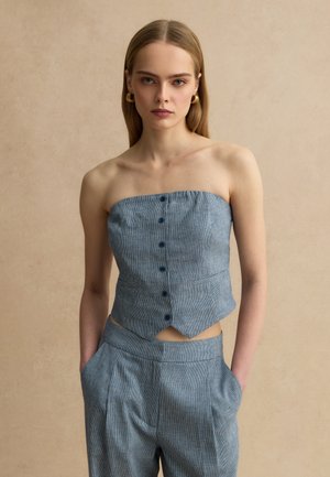 Junge Frau mit glattem blondem Haar, die goldene Creolen, ein trägerloses blaues Oberteil mit Knöpfen und passende hochtaillierte Hosen trägt, die Hände in den Taschen.