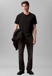 T-shirt preta de gola redonda, jeans cinza escuro de perna reta e sapatos pretos sem atacadores. Casaco preto pendurado no braço, com pouca publicidade na t-shirt.