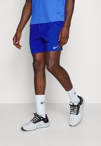 Pantalones cortos deportivos azules, zapatillas negras y blancas con un acento dorado, y calcetines blancos cortos con un logotipo, combinados con una camiseta deportiva azul.