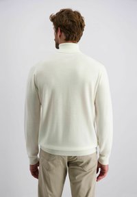 Weißer gestrickter Pullover mit hohem Kragen, mit gerippten Bündchen und Saum, über hellbeigen Hose getragen, von hinten gesehen.