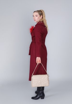 Cappotto di lana borgogna con cintura e decorazione in piume rosse, abbinato a una borsa trapuntata color crema e stivaletti neri. Tessuto liscio.
