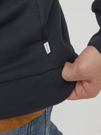 Maglione blu navy con polsini e orlo a costine, caratterizzato da un etichetta bianca con scritto "JACK & JONES." Mano che solleva leggermente l'orlo.