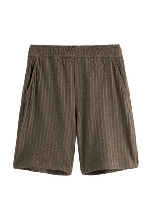 Dreng står iført en brun tekstureret skjorte med knapper og korte ærmer samt matchende knælange shorts med hvide sokker og sorte sneakers.