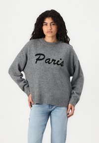 Fată tânără cu păr creț, purtând un pulover gri cu inscripția "Paris" în litere negre și blugi albaștri deschiși, stând cu o mână pe șold.