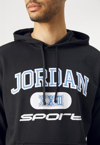 Felpa nera con cappuccio con la scritta "JORDAN" e "sport" stampate in bianco e azzurro chiaro. Il materiale ha una texture liscia e include un cappuccio con cordoncino.