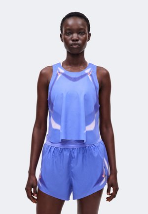 Vrouwelijke atleet draagt een blauw mouwloos sporttopje en bijpassende korte broek met subtiele oranje Nike-logo's, staand tegen een effen lichte achtergrond.