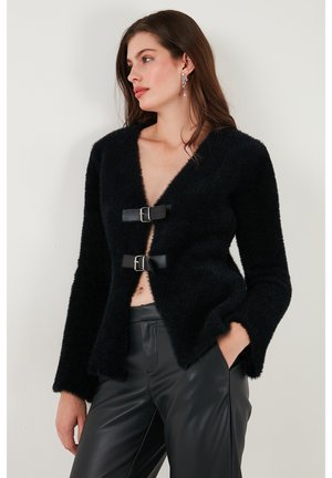 Zwarte fuzzy cardigan met een diepe V-hals, voorzien van twee frontgespen. Het heeft lange mouwen en een ontspannen, open ontwerp. Gekoeld met aansluitende zwarte broek.
