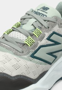 Chaussure de sport gris clair dotée d'une tige en mesh respirant avec des accents verts, des détails texturés et un logo vert foncé bien visible.