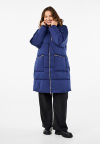 Marineblauwe pufferjas met capuchon, voorzien van een rits aan de voorkant, twee ritszakken en een gequilt patroon. Gemaakt over zwarte broeken en stevige zwarte laarzen.
