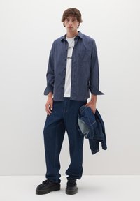Camicia a quadri blu navy e bianca con bottoni, t-shirt bianca con grafica, pantaloni in denim blu larghi, scarpe con tacco nero, mentre tiene una giacca di jeans.