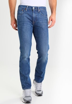 Homme portant un jean en denim bleu et des baskets blanc-gris, debout devant un fond clair uni.