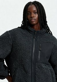 Giacca in pile nero sfocato con zip frontale, collo alto e tasca sul petto con zip. Presenta pannelli laterali lisci e ampie tasche laterali.