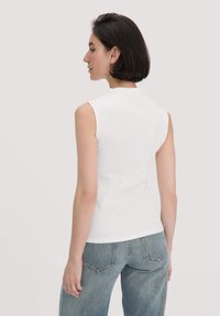 Witte mouwloze top met een geribbelde textuur, met een hoge halslijn. Gecombineerd met lichtblauwe spijkerbroek, weergegeven vanuit de achterkant.