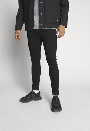 Homme portant un pantalon noir slim, des baskets noires avec des chaussettes blanches, une chemise grise et une veste noire, debout devant un fond uni.