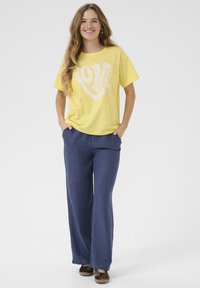 Kollane puuvillane t-särk valge südamegraafikaga, kombineerituna siniste linane-sinise laiade pükstega ja pruunid leopardimustriga tossudega.