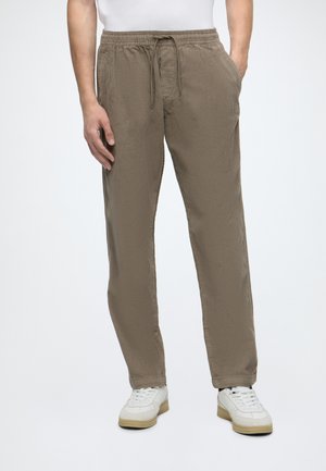 Homme portant un pantalon en velours côtelé marron clair avec taille élastique et cordon de serrage, associé à des baskets blanches et un haut blanc.