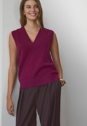 Magenta sleeveless sweater with a V-neck and ribbed texture. Paired with high-waisted, dark brown pleated trousers.