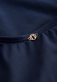 Primer plano de una tela azul oscuro con textura fina de malla y un pequeño emblema redondo de oro cosido en la costura.