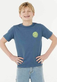 T-shirt en coton bleu à manches courtes et col rond, avec un graphisme circulaire vert sur la poitrine gauche. Porté avec un short en denim clair.