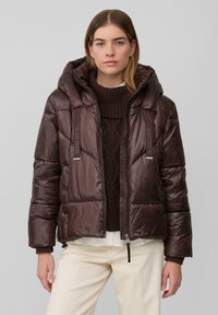 Braune Steppjacke mit glänzendem Finish, angebrachtem Kapuzen, Frontreißverschluss und Kordelzügen mit silbernen Akzenten. Trägt darüber einen dunkel gestrickten Pullover.