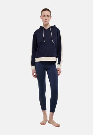 Jeune femme debout pieds nus, portant un sweat à capuche bleu marine avec des rayures crème et des leggings assortis bleu marine, sur un fond uni.