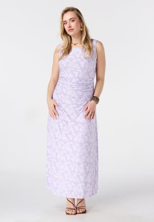 Vrouw staat en draagt een mouwloze lavendel bloemige maxijurk, gouden oorbellen, ketting, armband en bruine sandalen met bandjes.