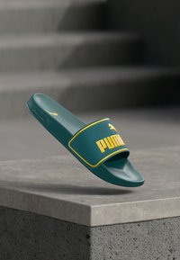 Πράσινο σανδάλι Puma slide με κίτρινο λογότυπο και περίγραμμα, τοποθετημένο πάνω από ένα σκυρόδεμα σκαλοπάτι με θολές σκάλες στο φόντο.