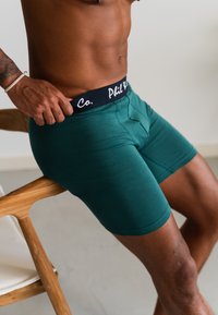 Gröna bomullshorts med svart midjeband som har texten "Phil Co." Modellen har ett silverarmband på handleden och sitter på en trästol.
