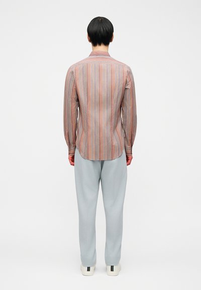 Άτομο που στέκεται πλάτη, φοράει μακρυμάνικο πουκάμισο με κάθετες πολύχρωμες ρίγες, ανοιχτό γκρι παντελόνι και λευκά αθλητικά παπούτσια με μαύρες ρίγες.