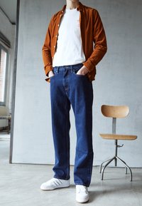 Brun skjorta med knappar över en vit t-shirt, kombinerad med blå jeans och vita sneakers, stående i en minimalistisk inomhusmiljö.