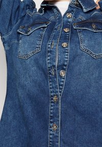 Lähikuva sinisestä denimpusakasta, jossa on hopeiset napit ja kaksi rintataskua, käytössä valkoisen paidan päällä.