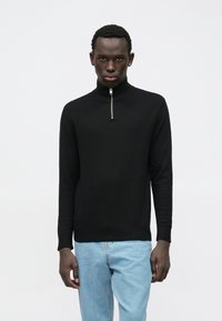 Moški nosi črn pulover z dolgimi rokavi in kratkim sprednjim zadrgo ter svetlo modre kavbojke, na enobarvni beli ozadju.