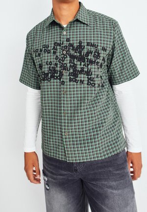 Kurzärmeliges grün-schwarz kariertes Hemd mit schwarzer Textgrafik, getragen über einem langärmeligen weißen Hemd mit dunklen, abgenutzten Jeans.