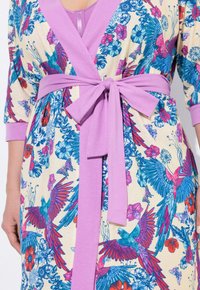 Vestido envolvente floral con una base crema y vibrantes patrones de pájaros azules y rojos. Cinturón morado que acentúa la cintura y mangas de 3/4.