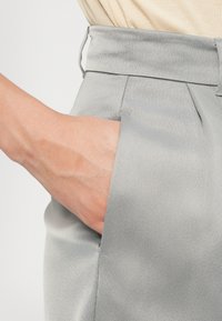 Pantalons gris clair avec une texture lisse, présentant une taille haute et des poches latérales. Le tissu semble légèrement réfléchissant.