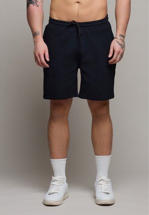 Mężczyzna w czarnych, teksturowanych szortach, białych stopkach i białych sneakersach, stojący na jednolitym tle.