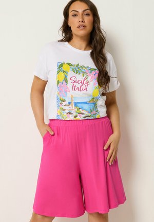 Kvinde med langt brunt hår iført en hvid Sicily Italia grafisk t-shirt og lyserøde vidde shorts med hænderne i lommerne.