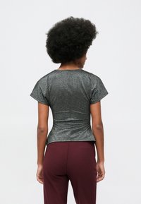 Žena s prirodnom afro frizurom nosi usku metalik sivu majicu kratkih rukava i burgundske hlače, prikazana sa leđa.