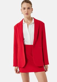 Rød blazer med et struktureret design, der har to forlommer og en knap. Sammen med en matchende rød nederdel og en hvid skjorte.