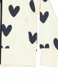 En creamfärgad zip-up hoodie i bomull med stora marinblå hjärtan, framficka och ribbade ärmslut. Har varumärkets etikett vid sidsömmen.